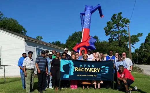 Group of people posing in front of APAA banner at the Association of Persons Affected by Addiction detox center in Dallas.
